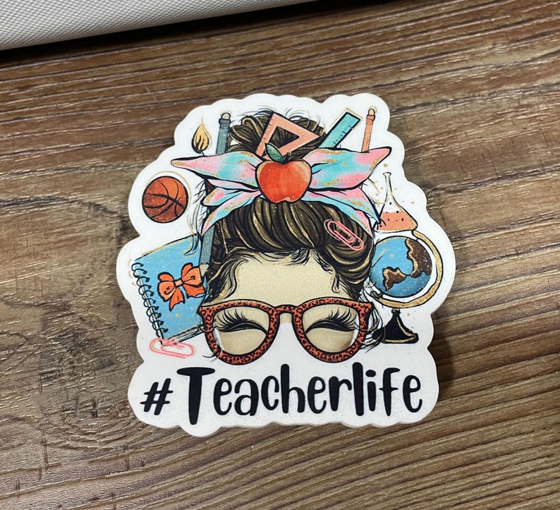 May include: White sticker with a cartoon illustration of a woman with a messy bun, wearing glasses and surrounded by school supplies. The text "#Teacherlife" is below the illustration.