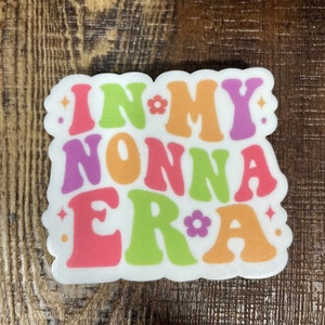 May include: White sticker with the text "In My Nonna Era" in colorful retro font with flowers and sparkles.