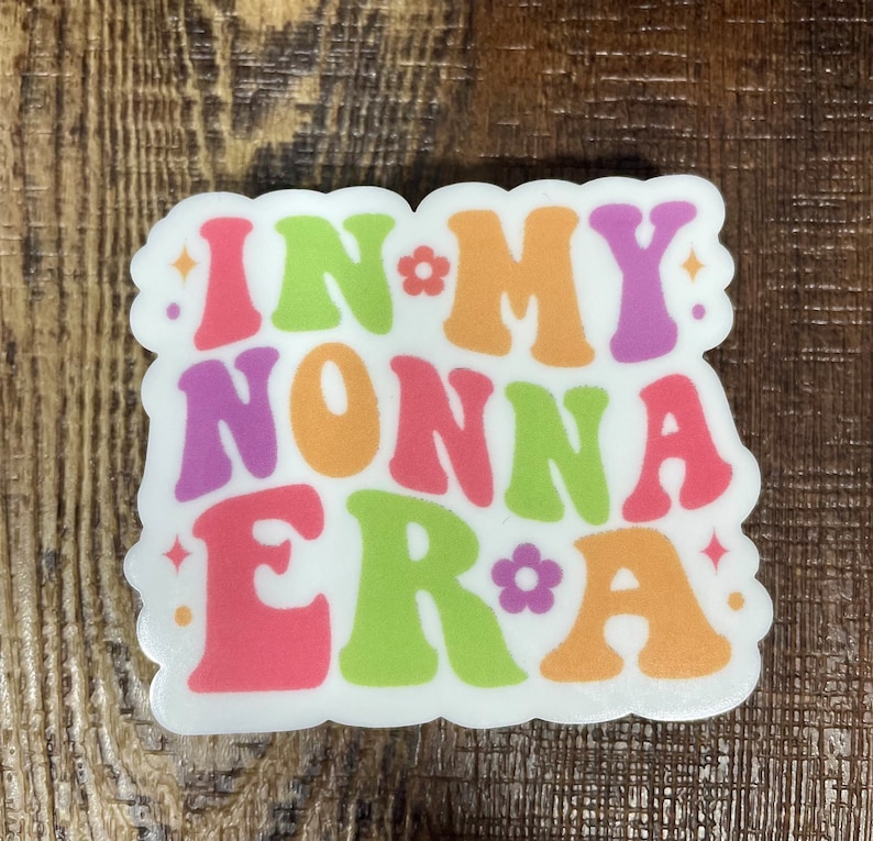 May include: White sticker with the text "In My Nonna Era" in colorful retro font with flowers and sparkles.