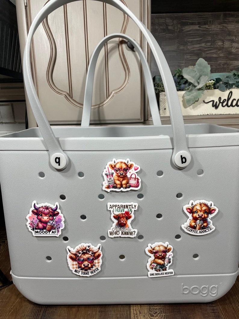 以下が含まれることがあります： 底に「bogg」という文字が印刷された灰色のトートバッグ。バッグには、ハイランド牛のカラフルなステッカーがいくつか貼られており、それぞれに「Moody AF」、「どうやら私は態度を持っているらしい、誰が知っていたの？」、「コーヒー中毒」、「今日はダメよ、ヒファー」、「ワン・ブージー・ヒファー」などの言葉が書かれている。