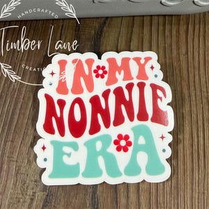 May include: A white sticker with the text "IN MY NONNIE ERA" in red, coral, and teal colors. The sticker has a retro font and floral accents. The sticker is on a wooden surface.