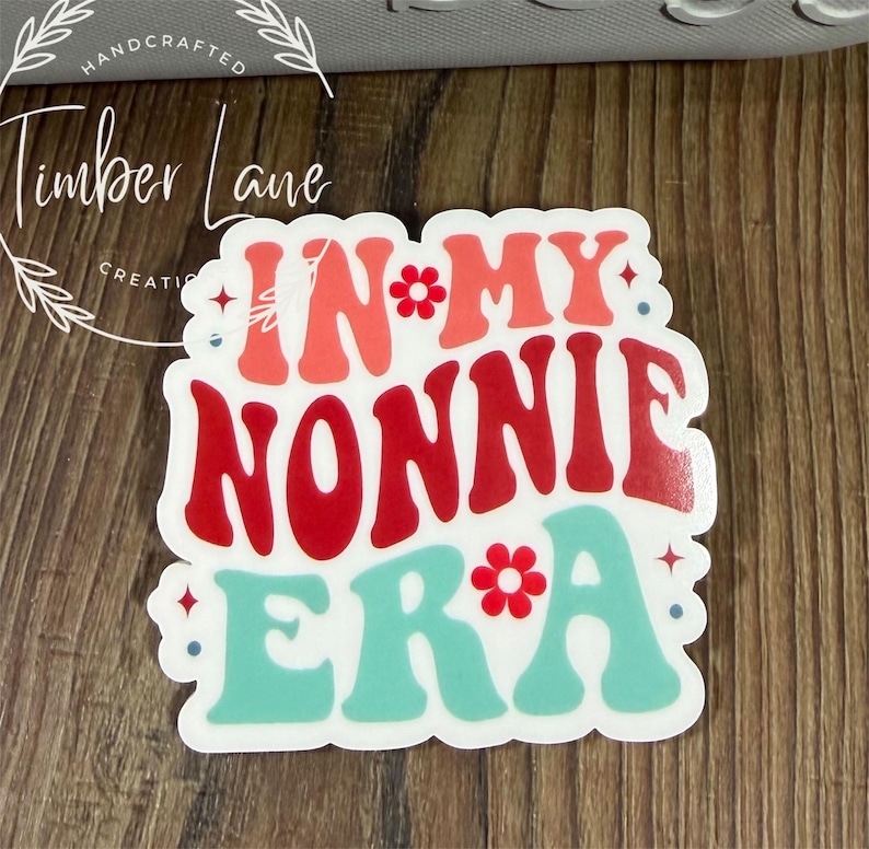 May include: A white sticker with the text "IN MY NONNIE ERA" in red, coral, and teal colors. The sticker has a retro font and floral accents. The sticker is on a wooden surface.