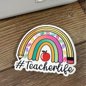 May include: A white sticker with a rainbow, a red apple, a pencil, and paper clips. The text "#Teacherlife" is written below the rainbow.