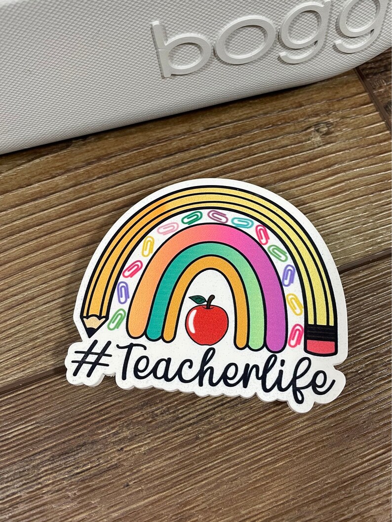 May include: A white sticker with a rainbow, a red apple, a pencil, and paper clips. The text "#Teacherlife" is written below the rainbow.