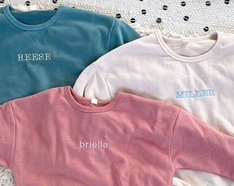 Könnte beinhalten: Drei langärmelige Sweatshirts in verschiedenen Farben: Türkis, Beige und Rosa. Jedes Sweatshirt hat einen anderen Namen auf der Vorderseite gestickt: Reese, Miller und Briella.