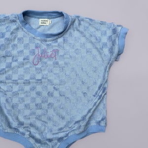 May include: A light blue baby onesie with a checkered pattern and the word "Juliet" embroidered on the front. The onesie has a crew neck and short sleeves.