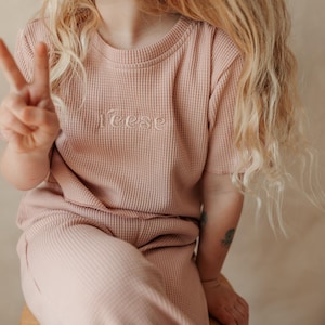 May include: A young person wearing a pink waffle knit top and matching pants. The top has the word "Reese" embroidered on the front.
