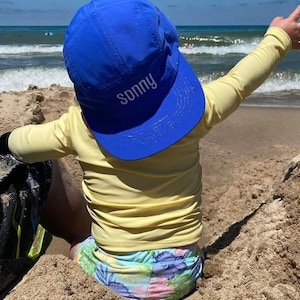 May include: A child wearing a yellow long-sleeved shirt and a blue baseball cap with the name "sonny" embroidered on it. The child is sitting in the sand on a beach.