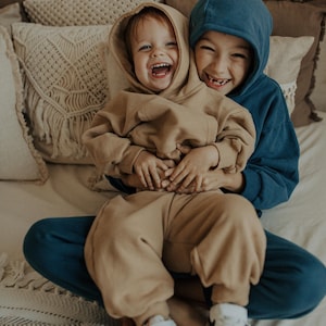 May include: Two children wearing matching hooded sweatshirts and sweatpants, one in tan and the other in blue. They are sitting together, smiling and laughing. The children are wearing white sneakers.
