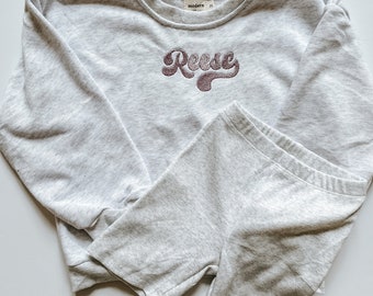 May include: A gray sweatshirt with the word "Reese" embroidered in a retro font on the front. The sweatshirt is paired with matching gray shorts.