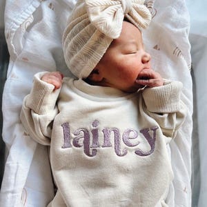 May include: A newborn baby wearing a cream-colored  sweatshirt with the name "Lainey" embroidered in purple. The baby is wearing a cream-colored turban with a large bow on top.