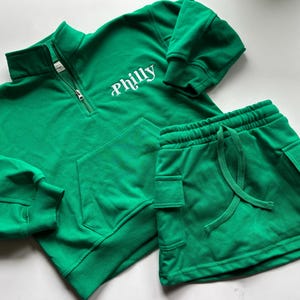 May include: A bright green two-piece outfit featuring a quarter-zip sweatshirt and matching shorts. The sweatshirt has the word "Philly" in white script. The shorts have a drawstring waist and cargo pockets. The set is made of a soft, comfortable material.