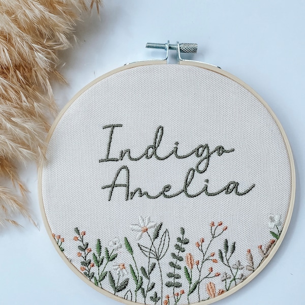 以下が含まれることがあります： 「Indigo Amelia」という名前が濃い緑色の筆記体で刺繍されたフープアート。オフホワイトの生地は木のフープで縁取られ、下端には白いヒナギクや緑の葉を含む花の刺繍デザインが施されています。