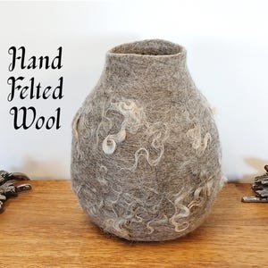May include: A gray hand-felted wool vase with white wool accents. The vase is round and has a wide opening at the top. "Hand Felted Wool" is written in black text on a white background.