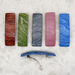 May include: Five rectangular hair clips with marbled patterns in shades of copper, green, gray, pink, and blue, arranged in a row. Below them, a single blue marbled hair clip is shown from the side, revealing its metal spring-loaded clasp mechanism.