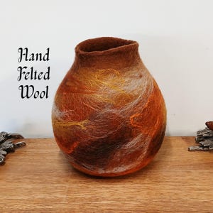 May include: A hand-felted wool vase with a rounded body and a flared opening. The vase features a blend of warm colors, including shades of brown, orange, and gold, with wisps of white. The text "Hand Felted Wool" is on the left.
