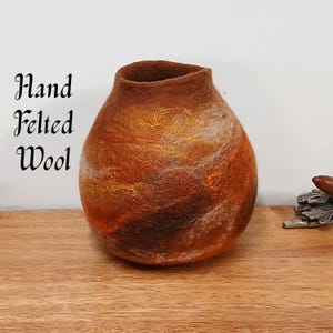 May include: A hand-felted wool vase in shades of brown, orange, and gold. The vase has a rounded body and a wide opening. The text "Hand Felted Wool" is on the left side of the vase.