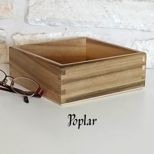 May include: A square wooden box with a light brown finish. The box is made of poplar wood and has a natural wood grain. The box is empty and has a smooth surface.