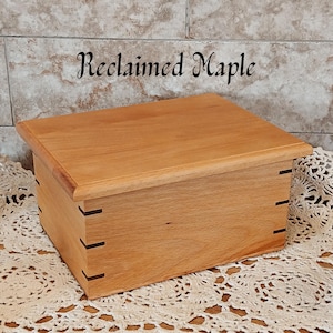 May include: A rectangular wooden box with a light brown finish. The box has a lid and is constructed with dovetail joints. The text "Reclaimed Maple" is above the box. The box is sitting on a white lace doily.