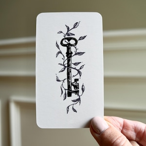 May include: A rectangular white card featuring a black ink illustration of an antique skeleton key entwined with delicate, leafy vines. The key is positioned vertically in the center of the card, showcasing a detailed bow, shaft, and bit.