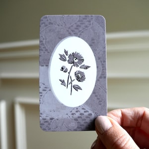 May include: A rectangular card with rounded corners featuring a grey lace-patterned background. In the center, a white oval contains a dark grey illustration of two blooming flowers with leaves and a small bud on a single stem, centered vertically.