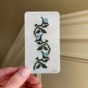 May include: A cross-stitched card featuring a vertical floral design in shades of green and blue. The design is on a white background. The card is rectangular with rounded corners. The artist's signature is in the bottom right corner.
