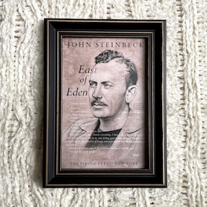 May include: A black framed print of John Steinbeck, the author of East of Eden. The print features a black and white illustration of Steinbeck with a quote from the book. The print is on a beige background with black text.