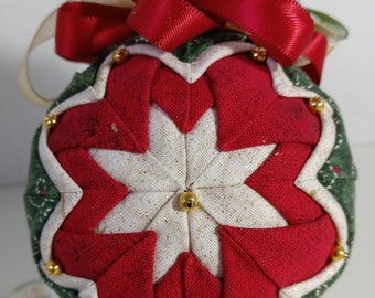 May include: A handcrafted Christmas ornament with a starburst design. The ornament features red and white fabric petals, accented with gold beads. A red ribbon and gold bow adorn the top, creating a festive holiday decoration.