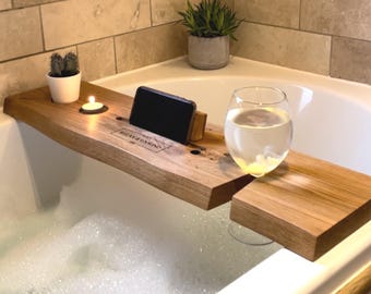May include: A rectangular wooden bathtub tray with a natural wood grain finish. The tray features a cutout for a smartphone, a circular holder for a tea light candle, and a slot for a wine glass. The text "RELAX & UNWIND" is engraved on the surface.