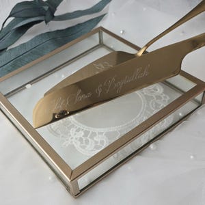 May include: A gold-coloured cake server and knife set, engraved with text, presented in a glass box with a gold frame. The set is placed on a white lace doily. The server reads "Elif Sena & Beytullah".