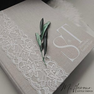 Peut inclure: Un livre beige recouvert de lin avec une bordure en dentelle blanche et une branche de feuilles vertes. Le livre porte les initiales "S & T" et les mots "Stammbuch Familie Meier" imprimés en blanc.