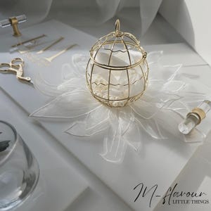 Op de afbeelding: Een gouden draadkooi met witte parels en een wit lint erin, die op een wit kantonderlegje ligt. De kooi staat op een wit oppervlak met de tekst "M-Havour LITTLE THINGS" in zwart.