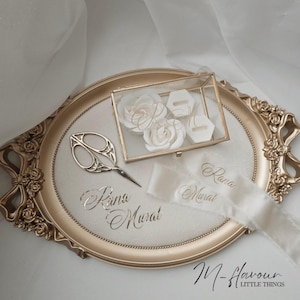 Könnte beinhalten: Ein goldfarbenes, ovales Tablett mit floralen Details hält eine Glas-Ringbox, weiße Rosen und eine Schere. Das Tablett zeigt auch ein weißes Band mit den Namen "Rana Murat" und dem Text "M-flavour LITTLE THINGS".