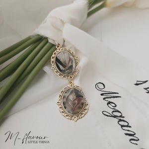 Könnte beinhalten: Doppelseitiger Anhänger in Goldton mit einem Rahmen im Vintage-Stil. Der Anhänger zeigt zwei ovale Bilder mit einem floralen Design. Der Anhänger hängt an einem weißen Band mit dem aufgestickten Text "Megan".