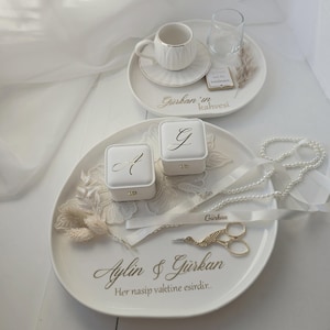 Op de afbeelding: Twee witte keramische trays met gouden letters en decoratieve items. Eén tray bevat een koffiekopje en een glas. De andere bevat twee ringdozen, een parelketting en een schaar. De tekst luidt "Aylin & Gürkan".