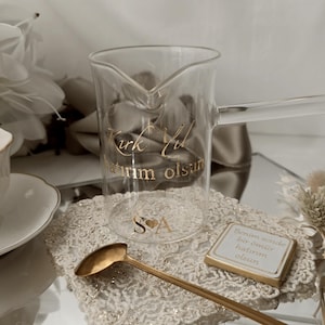 Puede incluir: Una cafetera de cristal transparente con un asa larga y un pico vertedor. La olla tiene letras doradas que dicen "Kırk Yıl Hatırım Olsun" y las iniciales "S♥A". También se ven una cuchara dorada y una pequeña tableta de chocolate.