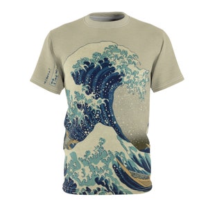 May include: A beige t-shirt with a print of "The Great Wave off Kanagawa" by Katsushika Hokusai. The print features a large blue wave with a white crest and a smaller wave in the foreground. There are three boats with people in them in the foreground of the print. The Japanese text on the shirt reads "神奈川沖浪裏" which translates to "The Great Wave off Kanagawa".