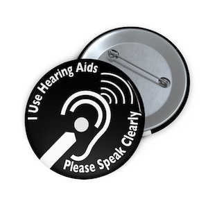 May include: Black and white button with the text "I Use Hearing Aids Please Speak Clearly" and a stylized image of an ear with a sound wave.