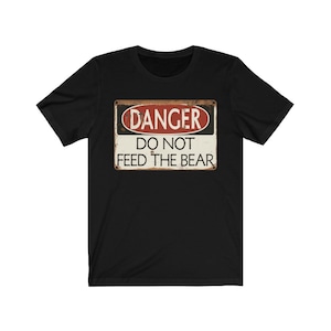 May include: Black t-shirt featuring a vintage-style warning sign that reads "DANGER DO NOT FEED THE BEAR".