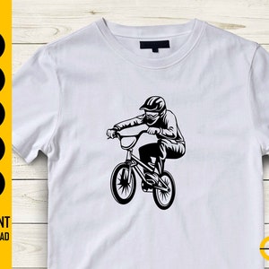 Op de afbeelding: Wit t-shirt met een zwarte afbeelding van een BMX-fietser in actie. Het ontwerp toont een helm en een bril. De linkerkant van het shirt heeft de tekst "SVG, DXF, PNG, EPS, AI" en "DIRECT DOWNLOADEN".