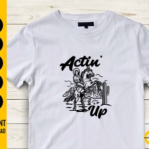 May include: White t-shirt featuring a black graphic of a woman riding a horse, with the words "Actin' Up" in a stylized font. The design includes desert scenery and cactus. The shirt is a casual wear item.