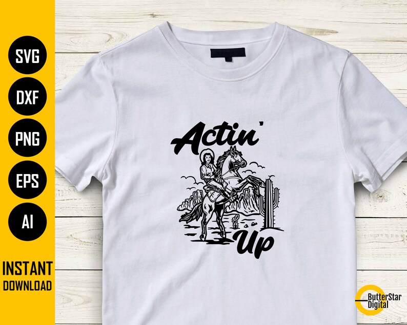 May include: White t-shirt featuring a black graphic of a woman riding a horse, with the words "Actin' Up" in a stylized font. The design includes desert scenery and cactus. The shirt is a casual wear item.