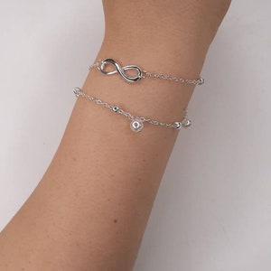 May include: Two silver bracelets on a person's wrist. The top bracelet features an infinity symbol charm. The bottom bracelet has small silver beads.