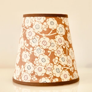 Peut inclure: Abat-jour conique orné d'un motif floral blanc sur fond marron. Les fleurs présentent des centres rouges et noirs. Les bords supérieur et inférieur sont bordés d'une bande marron foncé.