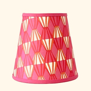 May include: A tapered drum lampshade featuring a vibrant pink background with a repeating geometric pattern of vertical, diamond-shaped segments in shades of orange, red, and white. The top and bottom edges are finished with a solid pink trim.