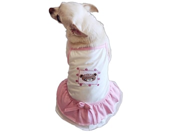 Peut inclure: Un chien blanc portant une robe rose et blanche avec un ours brun brodé sur le devant. La robe a un nœud rose au dos.