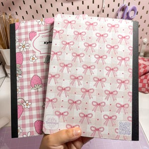 Może przedstawiać: Dwa zeszyty w kratkę z czarnymi grzbietami. Jeden zeszyt ma różowy wzór w kratkę z truskawkami i stokrotkami. Drugi ma różowy wzór kokardek na białym i różowym tle w kratkę. Zeszyty trzymane są w dłoni.