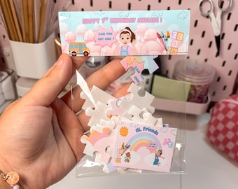 May include: A clear plastic bag filled with white puzzle pieces and a colorful card. The card features a rainbow, sun, and the text "Hi, Friends". The bag is topped with a header that reads "HAPPY 1ST BIRTHDAY AVALISE!" and "CAN YOU SAY ONE?"
