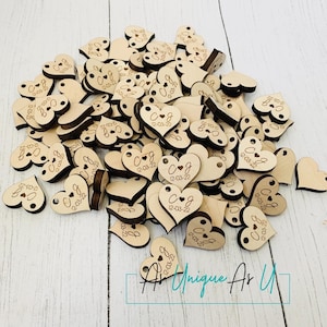 May include: A pile of wooden heart-shaped tags with the initials "CJ" and the date "12-03-22" engraved on them. The tags are scattered on a light wood surface.