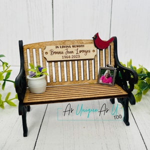 May include: A wooden bench with a personalized plaque that reads "In Loving Memory Bonnie Jean Limoges 1964-2023". A small red cardinal figurine sits on the back of the bench. A framed photo sits on the bench. The bench is black and brown and has a sign that reads "As Unique As You Too".